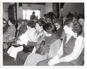B&W photo of a large group of people seated on rows of chairs in a hall. Many people are looking across to one side of the hall. In the background, a few people can be seen standing at the back of the hall. There is a large set of windows, partially obscured by the figure of a person walking towards the back of the hall.