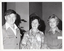 B&W medium close-up photo of three people posing for a photo inside a hall. Behind them, the brickwork of the building is visible on the interior walls. The silouhette of a tall potted plant is visible in front of one of the walls.