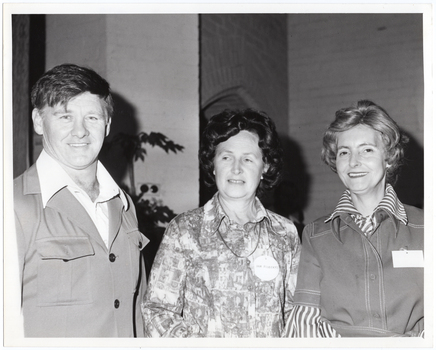 B&W medium close-up photo of three people posing for a photo inside a hall. Behind them, the brickwork of the building is visible on the interior walls. The silouhette of a tall potted plant is visible in front of one of the walls.
