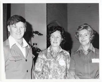 B&W medium close-up photo of three people posing for a photo inside a hall. Behind them, the brickwork of the building is visible on the interior walls. The silouhette of a tall potted plant is visible in front of one of the walls.