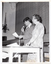 B&W photo of two people standing on the floor inside a hall, in front of a stage . There is a wooden table in front of them. The man is speaking into a microphone with a lead, which he is holding in one hand, while holding a piece of paper in his other hand. The woman is standing next to him, with one hand on the table. They are both depicted in profile, as they are facing away from the camera. There is a macrame plant holder hanging from the wall, with a potted plant inside it.