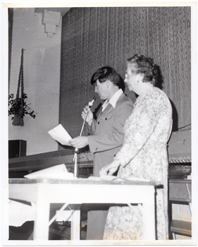 B&W photo of two people standing on the floor inside a hall, in front of a stage . There is a wooden table in front of them. The man is speaking into a microphone with a lead, which he is holding in one hand, while holding a piece of paper in his other hand. The woman is standing next to him, with one hand on the table. They are both depicted in profile, as they are facing away from the camera. There is a macrame plant holder hanging from the wall, with a potted plant inside it.