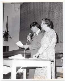 B&W photo of two people standing on the floor inside a hall, in front of a stage . There is a wooden table in front of them. The man is speaking into a microphone with a lead, which he is holding in one hand, while holding a piece of paper in his other hand. The woman is standing next to him, with one hand on the table. They are both depicted in profile, as they are facing away from the camera. There is a macrame plant holder hanging from the wall, with a potted plant inside it.