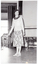 B&W photo of a woman standing on some wooden floorboards, smiling. Behind her, a large castor wheel at the base of a metal leg (which forms part of an unidentified item) is visible in the background. Various other unidentifiable objects are visible beyond an open concertina doorway beside the audience.