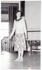 B&W photo of a woman standing on some wooden floorboards, smiling. Behind her, a large castor wheel at the base of a metal leg (which forms part of an unidentified item) is visible in the background. Various other unidentifiable objects are visible beyond an open concertina doorway beside the audience.
