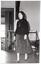 B&W photo of a woman walking on some wooden floorboards, smiling. Behind her, there are some clothes hanging on a short clothes rack. In the background, there is a large castor wheel at the base of a metal leg, (which forms part of an unidentified item). Behind her, on one side, there are various other unidentifiable objects beyond an open concertina door.