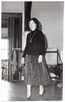 B&W photo of a woman walking on some wooden floorboards, smiling. Behind her, there are some clothes hanging on a short clothes rack. In the background, there is a large castor wheel at the base of a metal leg, (which forms part of an unidentified item). Behind her, on one side, there are various other unidentifiable objects beyond an open concertina door.