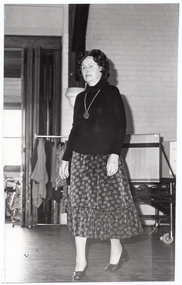B&W photo of a woman walking on some wooden floorboards, smiling. Behind her, there are some clothes hanging on a short clothes rack. In the background, there is a large castor wheel at the base of a metal leg, (which forms part of an unidentified item). Behind her, on one side, there are various other unidentifiable objects beyond an open concertina door.