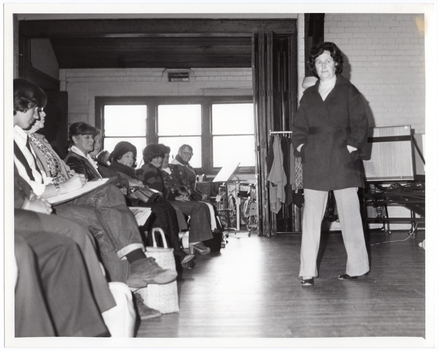 B&W photo of a woman walking on some wooden floorboards. She is modelling an outfit in front of a seated audience. Behind her, there are some clothes hanging on a short clothes rack. In the background, there is a large castor wheel at the base of a metal leg, (which forms part of an unidentified item). Behind her, on one side of the audience, there are various other unidentifiable objects beyond an open concertina door.