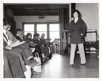 B&W photo of a woman walking on some wooden floorboards. She is modelling an outfit in front of a seated audience. Behind her, there are some clothes hanging on a short clothes rack. In the background, there is a large castor wheel at the base of a metal leg, (which forms part of an unidentified item). Behind her, on one side of the audience, there are various other unidentifiable objects beyond an open concertina door.