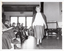 B&W photo of a woman walking on some wooden floorboards. She is modelling an outfit in front of a seated audience. Behind her, there are some clothes hanging on a short clothes rack. In the background, there is a large castor wheel at the base of a metal leg, (which forms part of an unidentified item). Behind her, on one side of the audience, there are various other unidentifiable objects beyond an open concertina door.