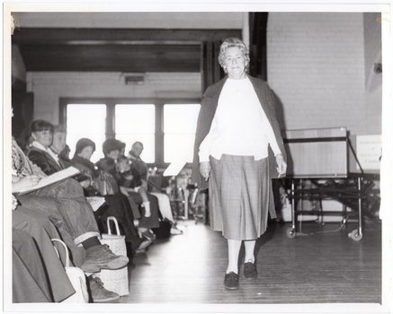 B&W photo of a woman walking on some wooden floorboards. She is modelling an outfit in front of a seated audience. Behind her, there are some clothes hanging on a short clothes rack. In the background, there is a large castor wheel at the base of a metal leg, (which forms part of an unidentified item). Behind her, on one side of the audience, there are various other unidentifiable objects beyond an open concertina door.