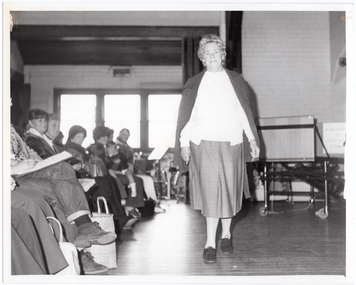 B&W photo of a woman walking on some wooden floorboards. She is modelling an outfit in front of a seated audience. Behind her, there are some clothes hanging on a short clothes rack. In the background, there is a large castor wheel at the base of a metal leg, (which forms part of an unidentified item). Behind her, on one side of the audience, there are various other unidentifiable objects beyond an open concertina door.