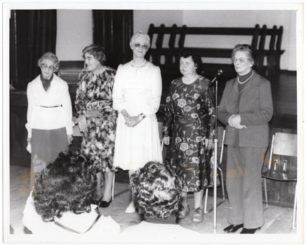 B&W photo of five women standing on the floor in front of a stage. The first four women are standing together in a row, while the fifth woman, on the far right, is speaking into a microphone on a floor stand. There is a row of chairs behind them. In the background, four wooden benches are stacked close to one another on the stage. In the foreground, there are two seated audience members who have been Photographed from behind.