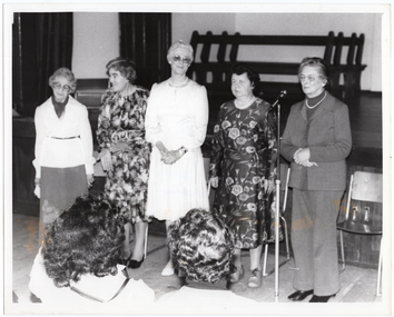 B&W photo of five women standing on the floor in front of a stage. The first four women are standing together in a row, while the fifth woman, on the far right, is speaking into a microphone on a floor stand. There is a row of chairs behind them. In the background, four wooden benches are stacked close to one another on the stage. In the foreground, there are two seated audience members who have been Photographed from behind.