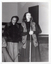B&W photo of a woman standing on the floor in front of a stage. She is speaking into a microphone on a stand. She is wearing a crocheted vest over a dark slong-sleeved top and a skirt. Standing behind her is a man, with another man (partially obscured) seated beside him. He is wearing a corduroy jacket and trousers. In the background, there are some wooden benches stacked close to one another on the stage.