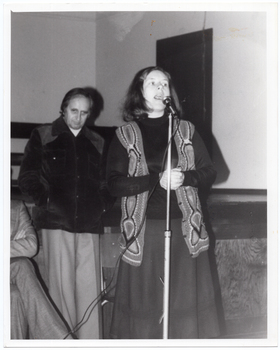 B&W photo of a woman standing on the floor in front of a stage. She is speaking into a microphone on a stand. She is wearing a crocheted vest over a dark slong-sleeved top and a skirt. Standing behind her is a man, with another man (partially obscured) seated beside him. He is wearing a corduroy jacket and trousers. In the background, there are some wooden benches stacked close to one another on the stage.