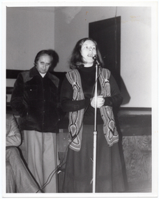 B&W photo of a woman standing on the floor in front of a stage. She is speaking into a microphone on a stand. She is wearing a crocheted vest over a dark slong-sleeved top and a skirt. Standing behind her is a man, with another man (partially obscured) seated beside him. He is wearing a corduroy jacket and trousers. In the background, there are some wooden benches stacked close to one another on the stage.