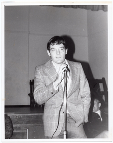 B&W photo of a man standing on the floor in front of a stage. He is speaking into a microphone on a stand. He is wearing eye glasses, and a checked jacket over a shirt and trousers. Behind him, there are two people (partially obscured) sitting on chairs. One of them is smiling broadly while turning his head to the side towards the other person. He is wearing a corduroy jacket and trousers. In the background, there are some wooden benches stacked close to one another on the stage.