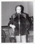B&W photo of a man standing on the floor in front of a stage. He is speaking into a microphone on a stand. He is wearing a corduroy jacket and trousers. Behind him, there are two other people (partially obscured) sitting on chairs. In the background, there are some wooden benches stacked close to one another on the stage.