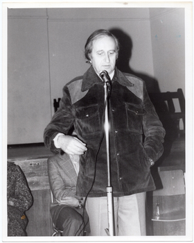 B&W photo of a man standing on the floor in front of a stage. He is speaking into a microphone on a stand. He is wearing a corduroy jacket and trousers. Behind him, there are two other people (partially obscured) sitting on chairs. In the background, there are some wooden benches stacked close to one another on the stage.