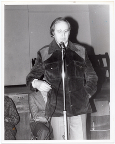 B&W photo of a man standing on the floor in front of a stage. He is speaking into a microphone on a stand. He is wearing a corduroy jacket and trousers. Behind him, there are two other people (partially obscured) sitting on chairs. In the background, there are some wooden benches stacked close to one another on the stage.