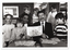 B&W photo of two men sitting at two small tables. One of the men is holding a brochure titled, "Children with Arthritis Family Support Group". There are several copies of the brochures on the tables. Sitting and standing around the men is a small group of children. Some are looking directly at the camera while others are looking at the brochure in the man's hands.