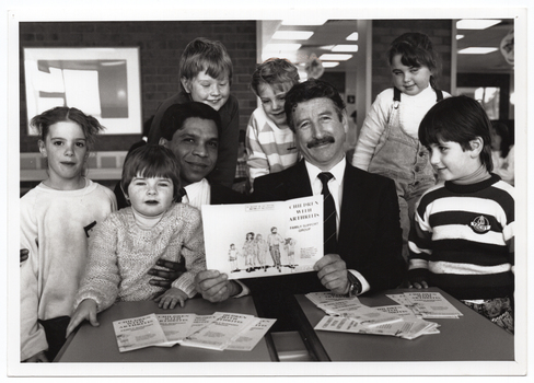 B&W photo of two men sitting at two small tables. One of the men is holding a brochure titled, "Children with Arthritis Family Support Group". There are several copies of the brochures on the tables. Sitting and standing around the men is a small group of children. Some are looking directly at the camera while others are looking at the brochure in the man's hands.