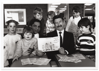 B&W photo of two men sitting at two small tables. One of the men is holding a brochure titled, "Children with Arthritis Family Support Group". There are several copies of the brochures on the tables. Sitting and standing around the men is a small group of children. Some are looking directly at the camera while others are looking at the brochure in the man's hands.