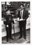 A man and a boy, both standing, pose for a photo in front of the counter of a fast food restaurant. The man (wearing a suit, shirt and tie), is carrying a tray of food and a drink. Next to him, the boy (wearing an "Australia" jumper with an Australian flag design within a large triangle) is using two walking sticks to support himself.