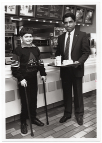 A man and a boy, both standing, pose for a photo in front of the counter of a fast food restaurant. The man (wearing a suit, shirt and tie), is carrying a tray of food and a drink. Next to him, the boy (wearing an "Australia" jumper with an Australian flag design within a large triangle) is using two walking sticks to support himself.