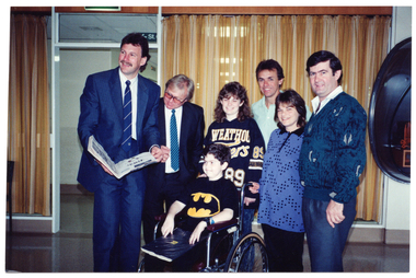 COL photo of a small group of people posing for a photo. A boy, who is a wheelchair user, is positioned at the front and centre of the group, while the others, all adults, are standing behind him. A woman has one hand on one of the wheelchair hand grips. A man on the left of the frame is holding a large open bound book, while the boy has a large closed book on his lap. Behind the group are some gold and beige striped curtains covering the large floor to ceiling windows. The windows are situated on either side of an open doorway. To the right of the frame, affixed to the wall, there is a smoky grey perspex telephone booth surrounding an orange public phone (partially obscured).