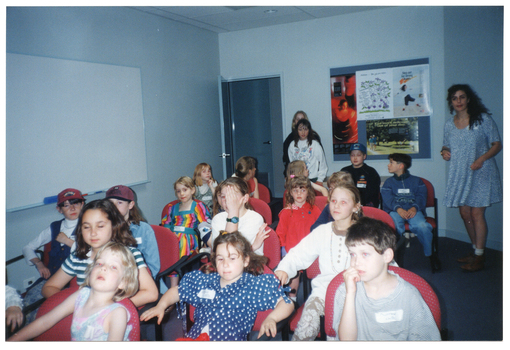 COL photo of a group of children seated on rows of chairs inside a room. In the background are two adults and a child standing near an open door and a display board, with posters, on the wall. There is a large whiteboard affixed to another wall.