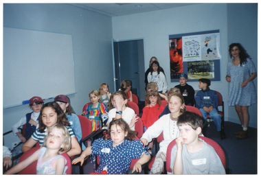 COL photo of a group of children seated on rows of chairs inside a room. In the background are two adults and a child standing near an open door and a display board, with posters, on the wall. There is a large whiteboard affixed to another wall.