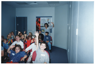 COL photo of a group of children seated on rows of chairs inside a room. Most of them have their hands and arms up in the air. In the background, there is a woman standing near an open door and a display board, with posters, on the wall. She also has her hands and arms up. Several closed bi-fold door panels are visible on the right-hand side of the frame.