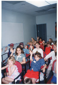 COL photo of a group of children seated on rows of chairs inside a room. Many of them are holding their hands up, some with two fingers depicting the "peace" sign. In the background, there is a whiteboard affixed to one wall, with an open door and a display board with posters (partially obscured) on the other wall.