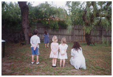 COL photo of a small group of children playing a game on a grassy area outdoors. A woman is crouching beside some of the children. The photo has been taken from behind them as they stand, facing the fence.
