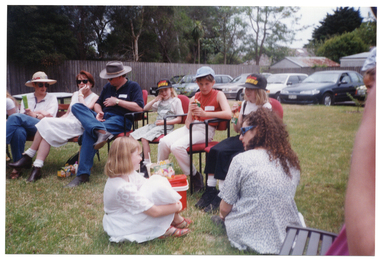 COL photo of a group of children and adults outdoors, either sitting on chairs or on the grass. There is an esky on the grass. In the background, in the carpark beyond the grassy area, there is a row of parked cars.