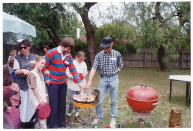 COL photo of a group of children and adults gathered on a grassy area outdoors, either sitting on chairs or standing. Two men are cooking various meats on a barbeque while while two girls look on. One of the girls has her arm wrapped around one of the men's arm. There is a second barbeque next to them, with the lid closed. Behind them, there is a woman holding a stack of round plastic trays. In the background, there are several other children and adults, standing or sitting under a large outdoor umbrella.