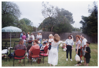 COL photo of a group of children and adults gathered on a grassy area outdoors. Some are sitting on chairs while others are standing. In the foreground, there is a woman holding a baby and another woman holding a couple of plastic bags full of bread. In the background, there are several people gathered around a barbeque. There is a large outdoor umbrella on a stand to the left, and a couple of tables with various miscellaneous items on them to the right.