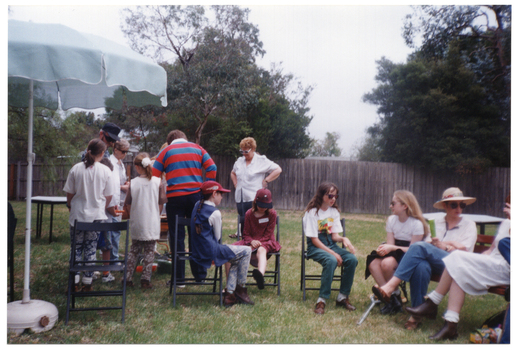 COL photo of a group of children and adults gathered on a grassy area outdoors. A small group of people is standing around a barbeque while others are seated nearby. To the left, there is a large outdoor umbrella on a stand.