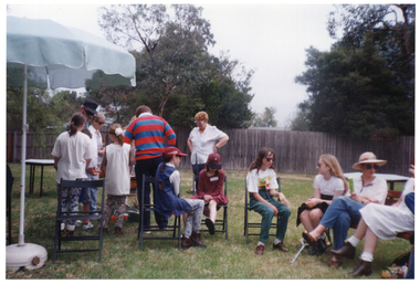COL photo of a group of children and adults gathered on a grassy area outdoors. A small group of people is standing around a barbeque while others are seated nearby. To the left, there is a large outdoor umbrella on a stand.