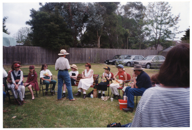 COL photo of a group of children and adults gathered on a grassy area outdoors. Most are sitting on chairs and a wooden bench while a couple of them are standing. In the background, beyond the grassy area, a row of parked cars is visible.