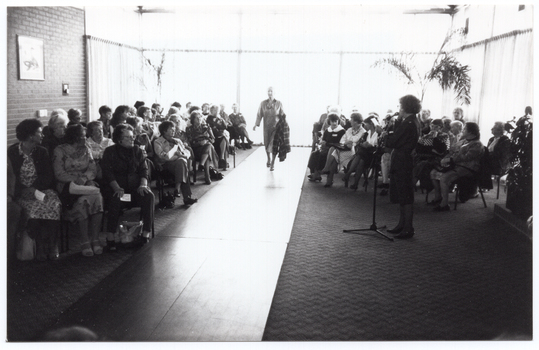 B&W photo of the interior of large reception room. A woman is speaking into a microphone on a stand while another woman models an outfit on a catwalk. The audience, seated on either side of the catwalk, looks on. Behind them are several large windows along two walls and part of another wall. The windows are covered in long sheer curtains to the floor.
