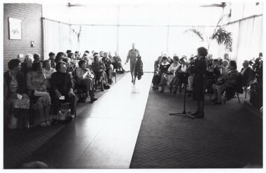 B&W photo of the interior of large reception room. A woman is speaking into a microphone on a stand while another woman models an outfit on a catwalk. The audience, seated on either side of the catwalk, looks on. Behind them are several large windows along two walls and part of another wall. The windows are covered in long sheer curtains to the floor.