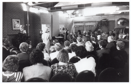 B&W photo of the interior of large reception room. A woman is speaking into a microphone on a stand while another woman models an outfit. She is surrounded by a large audience, who are seated in rows which curve around, extending beyond the end of the catwalk. Behind the woman at the microphone are two other women: one is seated behind a small table and the other is standing next to her. At the far end of the room, behind the audience, there is a clothing rack holding several coats on coathangers. There are three large potted plants in the room: one is situated in front of a brick fireplace and the other two are potted palms alongside another wall, near the clothing rack. The room consists of feature brick walls and there are no windows visible within the frame.