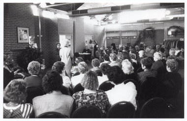 B&W photo of the interior of large reception room. A woman is speaking into a microphone on a stand while another woman models an outfit. She is surrounded by a large audience, who are seated in rows which curve around, extending beyond the end of the catwalk. Behind the woman at the microphone are two other women: one is seated behind a small table and the other is standing next to her. At the far end of the room, behind the audience, there is a clothing rack holding several coats on coathangers. There are three large potted plants in the room: one is situated in front of a brick fireplace and the other two are potted palms alongside another wall, near the clothing rack. The room consists of feature brick walls and there are no windows visible within the frame.