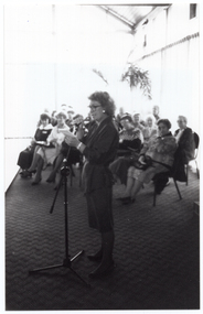 B&W photo of the interior of large reception room. A woman is speaking into a microphone on a stand while the audience, seated on one side of the catwalk, looks on. Behind them are several large windows along the walls, covered in long sheer curtains to the floor.