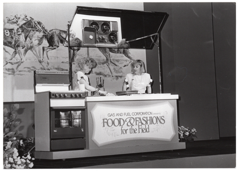 B&W photo of two women presenting a cooking demonstration on a stage. The women are standing behind a bench with an oven beside it. A sign on the front of the bench says "GAS AND FUEL CORPORATION presents FOOD & FASHIONS for the Field". Behind them is a large banner depicting a drawing of some jockeys riding racehorses, mid race.
