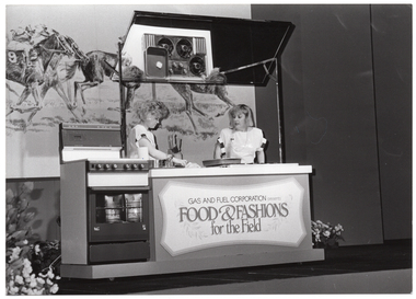 B&W photo of two women presenting a cooking demonstration on a stage. The women are standing behind a bench with an oven beside it. A sign on the front of the bench says "GAS AND FUEL CORPORATION presents FOOD & FASHIONS for the Field". Behind them is a large banner depicting a drawing of some jockeys riding racehorses, mid race.